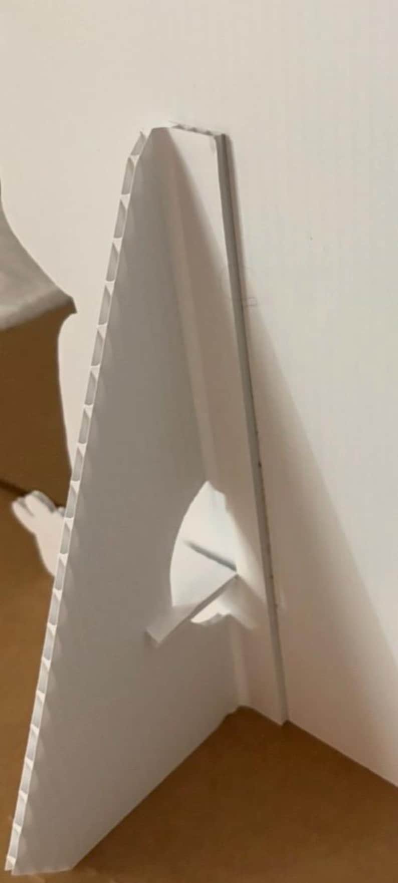 Op de afbeelding: Een witte golfkartonnen ezelrugsteun, ontworpen om rechtop te staan. De steun heeft een driehoekige vorm met een ingebouwde standaard voor stabiliteit. De randen hebben een geribbelde textuur.