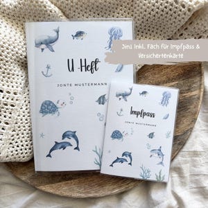 Könnte beinhalten: Ein weißes U-Heft und Impfpass Hüllenset mit Aquarellillustrationen blauer Meerestiere wie Wale, Delfine, eine Schildkröte und eine Qualle. Der Text lautet U-Heft, Impfpass, Jonte Mustermann sowie der Hinweis 3in1 inkl. Fach für Impfpass & Versichertenkarte.