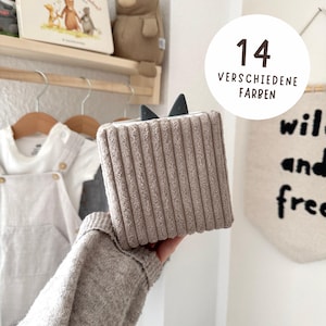 Könnte beinhalten: Eine taupefarbene Box mit Kordstruktur und dunkelgrauen Ohren wird in einer Hand gehalten. Der Text auf dem Bild lautet "14 VERSCHIEDENE FARBEN". Der Hintergrund umfasst Babykleidung und einen dekorativen Wandbehang.