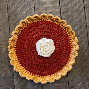 May include: A handmade crochet pie-shaped coaster. The coaster features a dark red center, a light tan crust, and a cream-colored flower in the center. The coaster is round and has a scalloped edge. The coaster is made of yarn.