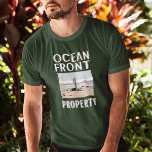 Puede incluir: Una camiseta verde oscuro con el texto "Ocean Front Property" en letras blancas. El dise&ntilde;o presenta una imagen de dibujos animados de un cactus en un paisaje des&eacute;rtico.