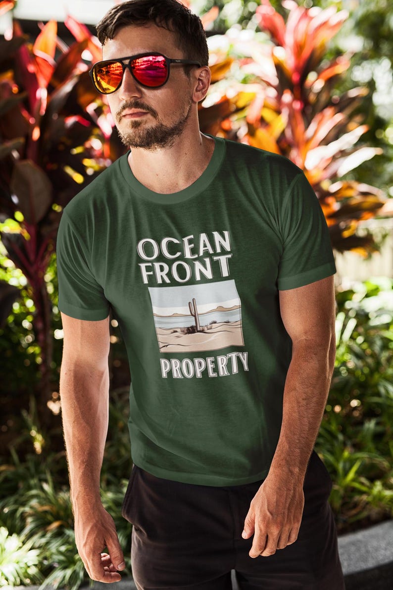 Puede incluir: Una camiseta verde oscuro con el texto "Ocean Front Property" en letras blancas. El dise&ntilde;o presenta una imagen de dibujos animados de un cactus en un paisaje des&eacute;rtico.