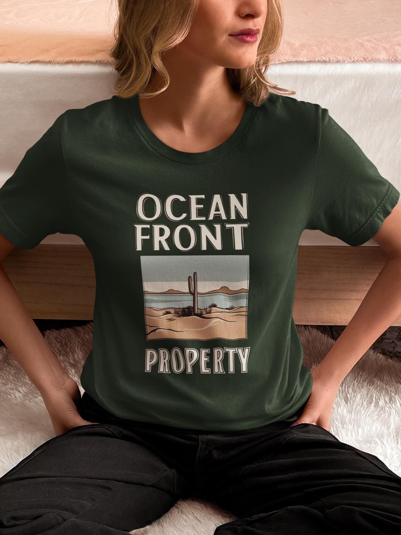 Puede incluir: Una camiseta verde oscuro con el texto "Ocean Front Property" y un gr&aacute;fico de un cactus y una escena de playa.