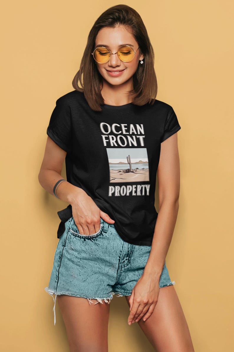 Puede incluir: Una camiseta negra con un dise&ntilde;o gr&aacute;fico de una escena del desierto con un cactus y el texto "Ocean Front Property".