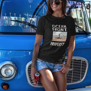 Puede incluir: Una mujer con una camiseta negra con el texto "Ocean Front Property" y un gr&aacute;fico de una escena de playa. Est&aacute; de pie frente a una furgoneta azul.