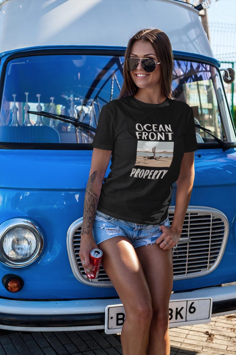 Puede incluir: Una mujer con una camiseta negra con el texto "Ocean Front Property" y un gr&aacute;fico de una escena de playa. Est&aacute; de pie frente a una furgoneta azul.