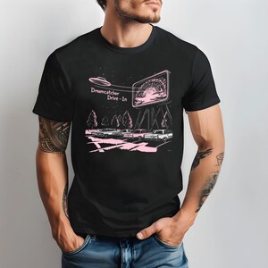 Puede incluir: Una camiseta negra con un gráfico rosa de un autocine con un OVNI volando por encima. El texto en el gráfico dice "Dreamcatcher Drive-In".