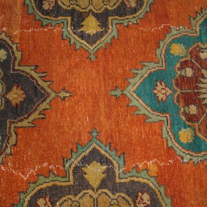 May include: Close-up of a distressed, vintage rug with an intricate floral pattern. The rug features a burnt orange background with teal, brown, and black floral medallions. The design includes a repeating pattern with a distressed, aged appearance.
