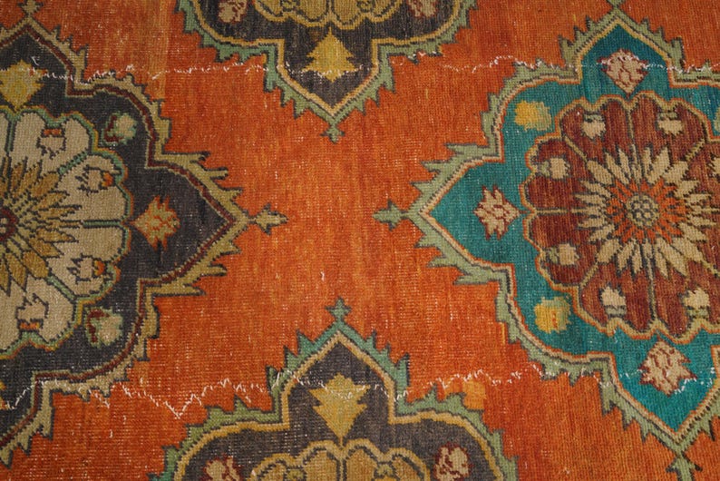 May include: Close-up of a distressed, vintage rug with an intricate floral pattern. The rug features a burnt orange background with teal, brown, and black floral medallions. The design includes a repeating pattern with a distressed, aged appearance.