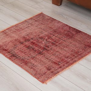 May include: A square area rug featuring a distressed, weathered texture in shades of deep red, burgundy, and faded pink. The rug has a low-pile construction with visible woven fibers and short, frayed fringe detailing along the edges.