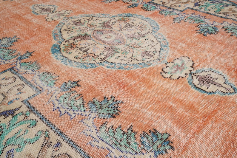 Pode incluir: Um tapete vintage com um fundo p&ecirc;ssego desbotado e um padr&atilde;o floral azul e verde. O tapete tem um aspecto desgastado e &eacute; feito de l&atilde;.