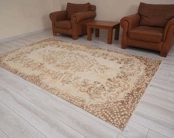 Op de afbeelding: Een rechthoekig, beige tapijt met een gedetailleerd bloemenpatroon en een bruine rand. Het tapijt ligt op een lichte houten vloer, met twee bruine leren fauteuils en een kleine houten tafel op de achtergrond.