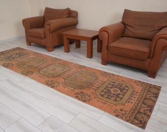 Op de afbeelding: Een patroon loper in oranje en bruine tinten, met een rechthoekig ontwerp. Het tapijt ligt op een lichte houten vloer, met twee bruine leren fauteuils en een kleine houten tafel op de achtergrond.
