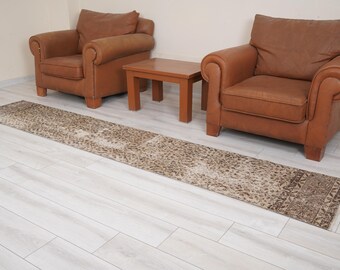 May include: A brown and beige patterned runner rug with a distressed look. The rug is placed on a light wood floor between two brown leather armchairs and a wooden coffee table.