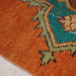 May include: Close-up of a section of a rug with a vibrant orange background. The rug features a detailed geometric design in teal, green, and brown. The texture of the rug is visible, showcasing the woven fibers.