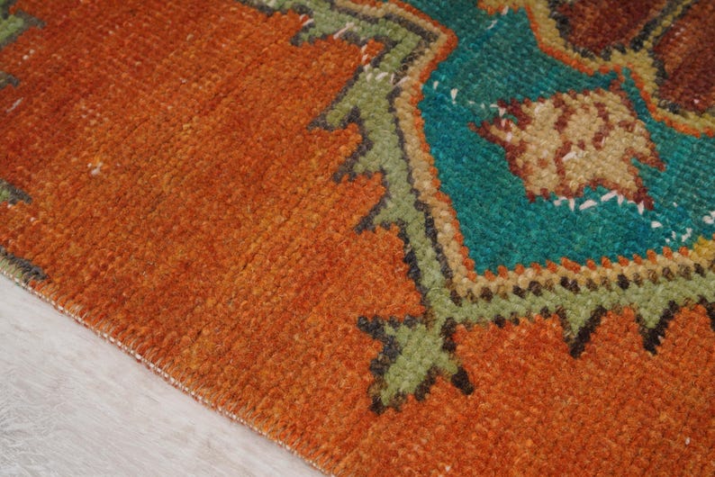 May include: Close-up of a section of a rug with a vibrant orange background. The rug features a detailed geometric design in teal, green, and brown. The texture of the rug is visible, showcasing the woven fibers.