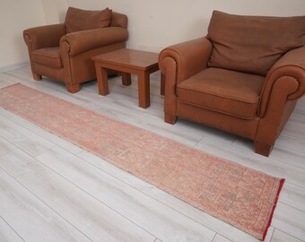 May include: A long, rectangular, vintage-style rug with a faded red and beige pattern. The rug is placed on a light-colored wooden floor, with two brown leather armchairs and a small wooden table in the background.