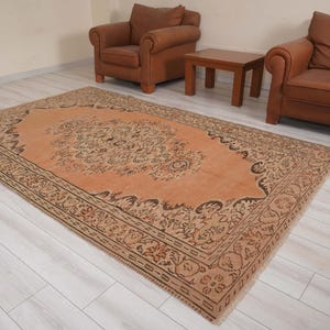 May include: An antique-style area rug with a central medallion design in shades of peach and brown. The rug features an ornate border and is placed on a light-colored wooden floor. The rug is rectangular and appears to be made of woven material.