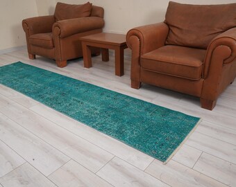 May include: A teal blue runner rug with a faded, distressed pattern. The rug is laid out on a light wood floor between two brown leather armchairs and a wooden coffee table.