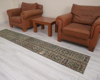 May include: A long, rectangular rug with a detailed geometric pattern in shades of green, brown, and beige. The rug is placed on a light-colored wooden floor, with two brown leather armchairs and a small wooden table in the background.