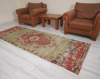 May include: A rectangular, distressed-look rug with a central medallion design in red and cream, surrounded by a faded green field. The rug is placed on a light wood floor, with two brown leather armchairs and a small wooden table in the background.