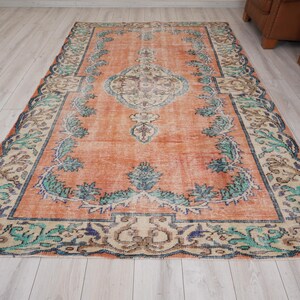 Pode incluir: Um tapete vintage com um fundo laranja desbotado e um padr&atilde;o floral em tons de verde, azul e bege. O tapete tem um aspeto desgastado e uma borda desfiada.