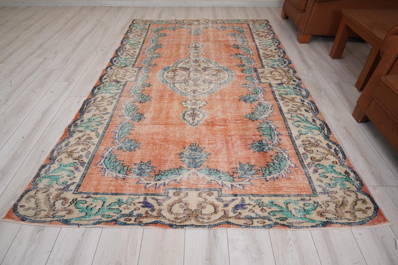 Pode incluir: Um tapete vintage com um fundo laranja desbotado e um padr&atilde;o floral em tons de verde, azul e bege. O tapete tem um aspeto desgastado e uma borda desfiada.