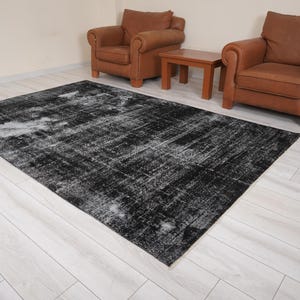 Pode incluir: Um tapete de área preto e branco desgastado com um visual desbotado e vintage. O tapete está colocado sobre um piso de madeira clara com duas poltronas de couro marrom e uma mesa de centro de madeira.
