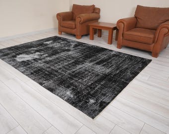 May include: A black and white distressed area rug with a faded, vintage look. The rug is laid out on a light wood floor with two brown leather armchairs and a wooden coffee table.