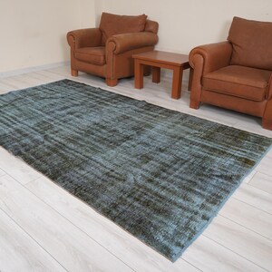 Pode incluir: Um tapete azul e verde com um padrão e um visual desgastado. O tapete está colocado sobre um piso de madeira clara entre duas poltronas de couro marrom e uma mesa de centro de madeira.