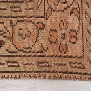 May include: Close-up of a section of a beige and brown patterned rug. The rug features geometric and floral designs in shades of brown and orange. The edge of the rug has a fringe.