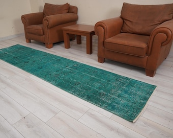 May include: A teal blue vintage runner rug with a faded pattern. The rug is laid out on a light wood floor in front of two brown leather armchairs.