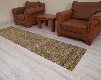 May include: A beige and green patterned runner rug with a fringe border. The rug is laid out on a light wood floor in front of two brown leather armchairs and a wooden coffee table.