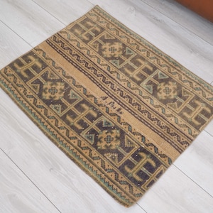 May include: A square, patterned rug with a beige background and dark brown and teal geometric designs. The rug has a central stripe and borders with repeating patterns. The rug is placed on a light-colored wooden floor.