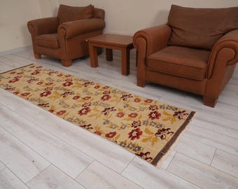 May include: A beige runner rug with a floral pattern in shades of red, yellow, and blue. The rug is laid out on a light wood floor between two brown leather armchairs.