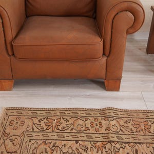 May include: A brown leather armchair with rolled arms and wooden legs sits near a small wooden side table. A patterned rug with a beige and brown design is in the foreground, adding warmth to the room.
