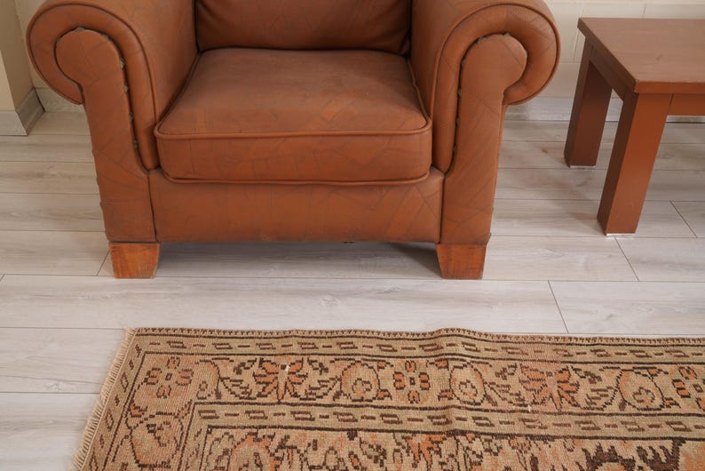 May include: A brown leather armchair with rolled arms and wooden legs sits near a small wooden side table. A patterned rug with a beige and brown design is in the foreground, adding warmth to the room.