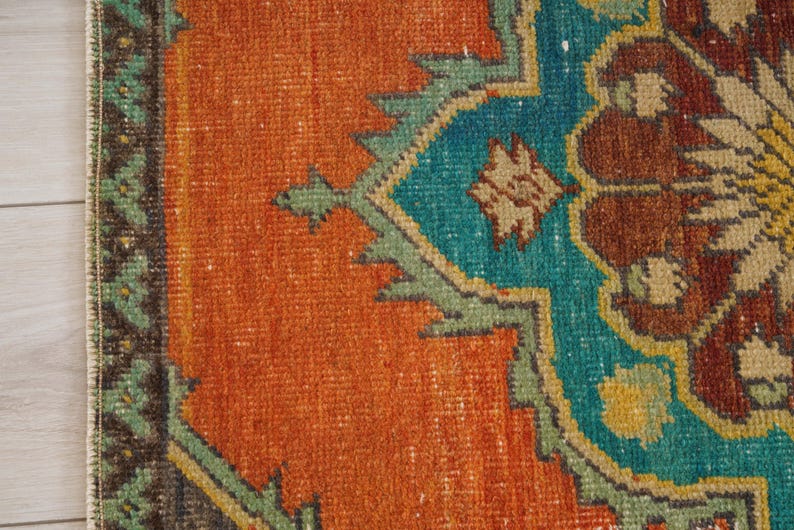 May include: Close-up of a section of a vintage rug with a detailed geometric pattern. The rug features a vibrant color palette of orange, teal, brown, and green. The design includes stylized floral and geometric shapes, creating a textured, antique aesthetic.