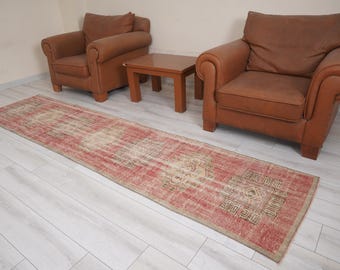 May include: A long, rectangular rug with a distressed, vintage appearance. The rug features a faded red and beige color palette with geometric patterns at one end. Two brown leather armchairs and a small wooden table are in the background.