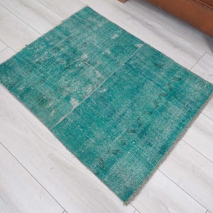 May include: A rectangular, distressed teal rug with a faded appearance. The rug is composed of several rectangular sections, creating a patchwork design. The rug's color is a vibrant teal with lighter, faded areas, giving it a vintage look. The rug is placed on a light-colored wooden floor.