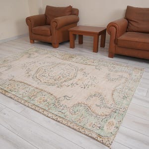 Pode incluir: Um tapete retangular de estilo vintage com um campo central creme e bordas verdes e florais. Duas poltronas de couro marrom e uma pequena mesa de madeira estão ao fundo, em um piso de madeira clara.
