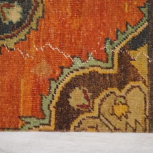 May include: Close-up of a section of a vintage rug with a distressed, textured appearance. The rug features a warm orange field with intricate patterns in brown, green, and yellow. The design has a traditional, oriental style.