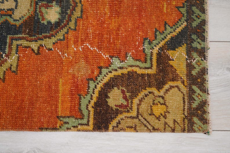 May include: Close-up of a section of a vintage rug with a distressed, textured appearance. The rug features a warm orange field with intricate patterns in brown, green, and yellow. The design has a traditional, oriental style.