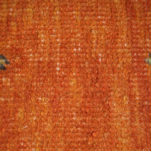 May include: Close-up of a textured orange rug with a repeating pattern. The rug has a dense, woven texture with a mix of orange, yellow, and white fibers. Two stylized green and black leaf-like shapes are visible.