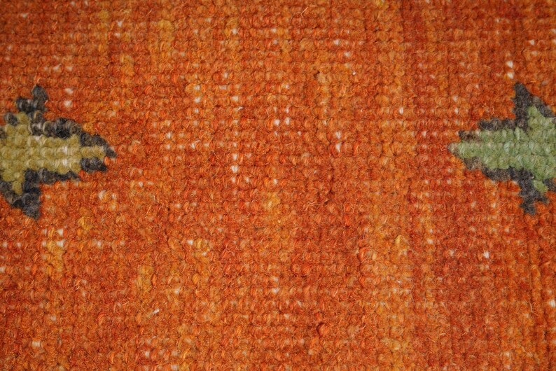 May include: Close-up of a textured orange rug with a repeating pattern. The rug has a dense, woven texture with a mix of orange, yellow, and white fibers. Two stylized green and black leaf-like shapes are visible.