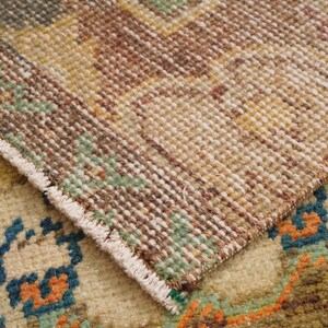 May include: Close-up of a section of a vintage rug with a detailed, textured weave. The rug features a palette of beige, brown, green, and orange hues. The edges of the rug are visible, showcasing the fringe and the intricate pattern.