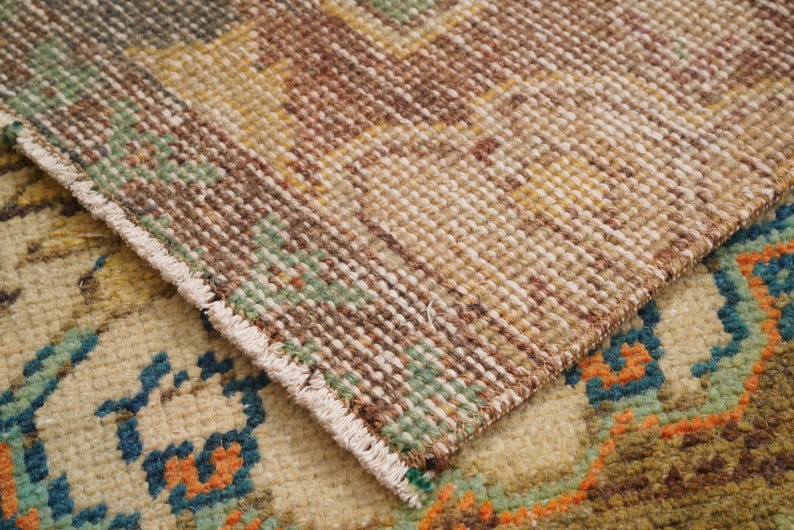 May include: Close-up of a section of a vintage rug with a detailed, textured weave. The rug features a palette of beige, brown, green, and orange hues. The edges of the rug are visible, showcasing the fringe and the intricate pattern.