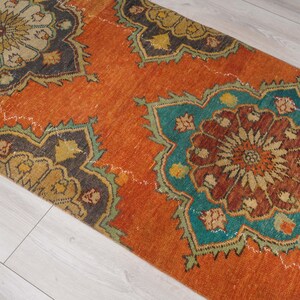 May include: An orange runner rug with a distressed, vintage look. The rug features large, repeating floral patterns in teal, brown, and gold. The rug's design is set against an orange background, creating a warm and inviting aesthetic.
