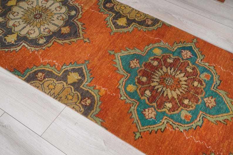 May include: An orange runner rug with a distressed, vintage look. The rug features large, repeating floral patterns in teal, brown, and gold. The rug's design is set against an orange background, creating a warm and inviting aesthetic.