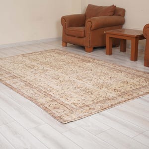 Pode incluir: Um tapete retangular com uma base de cor creme e padrões complexos de flores e vinhas em tons de castanho, bege e castanho claro. O tapete tem uma borda decorativa larga com motivos circulares repetidos e um design central denso.